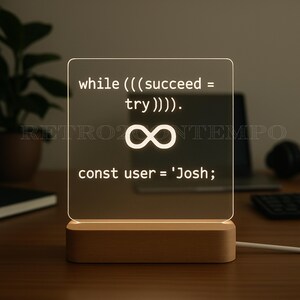 May include: A square, illuminated LED lamp with a wooden base. The lamp displays white text and an infinity symbol. The text reads: "while (((succeed = try)))). const user = 'Josh;" The lamp is plugged in and sits on a wooden surface.