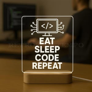 May include: A rectangular LED desk lamp with a wooden base. The lamp features a white graphic of a computer monitor with code symbols and the words "EAT SLEEP CODE REPEAT" in a bold, sans-serif font. The lamp is lit, and the background is blurred.