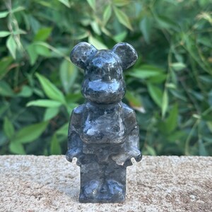 May include: A small, gray, stone figurine of a bear with large ears. The bear is standing on a gray, textured surface.