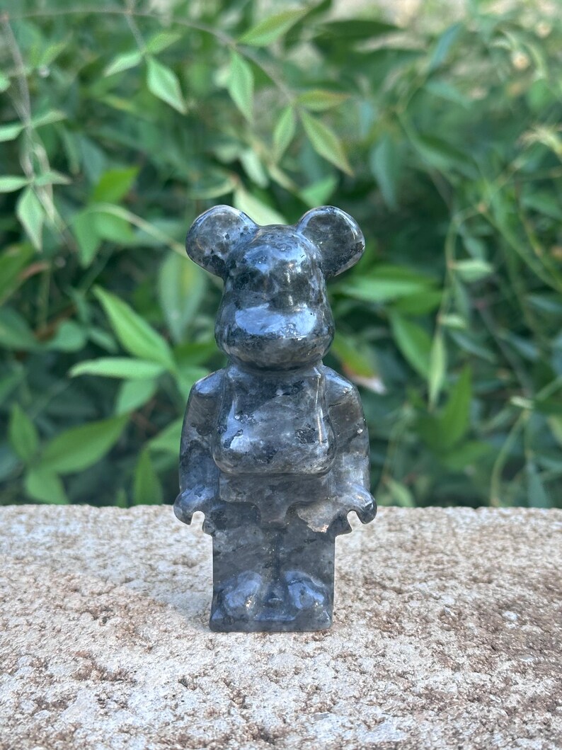May include: A small, gray, stone figurine of a bear with large ears. The bear is standing on a gray, textured surface.
