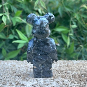 May include: A small, gray, stone figurine of a bear with a black and white marbled pattern. The bear is standing on a gray, textured surface.
