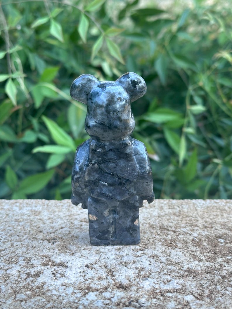 May include: A small, gray, stone figurine of a bear with a black and white marbled pattern. The bear is standing on a gray, textured surface.