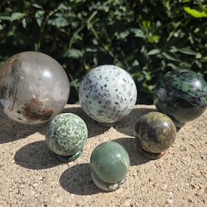 May include: A collection of six spherical stones of varying sizes and textures. The group includes a large clear quartz sphere with dark inclusions, a speckled light blue sphere, a dark green and purple sphere, and three smaller solid green spheres.