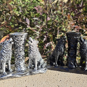May include: Two wolf figurines howling at a central pillar. One is silver and the other is black. The pillar has an eye design. The figurines are on a base that looks like a log. The background is out of focus.