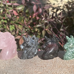 May include: Four carved unicorn figurines made of different types of stone. The unicorns are pink, black, dark purple, and green.
