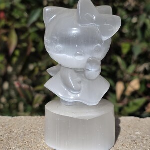 May include: A translucent white selenite figurine of a cat character, standing on a rectangular base. The cat has a bow on its head and is holding a small object. The figurine is set against a blurred green background.
