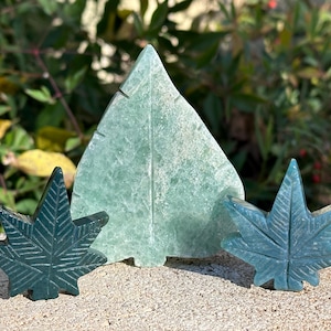 May include: Three carved stone sculptures in shades of green. A large, triangular leaf shape is flanked by two smaller, five-pointed leaf shapes. The sculptures are set on a textured, light-colored surface with a natural background.