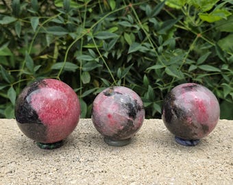 Puede incluir: Tres esferas de rodonita pulida que presentan una mezcla de inclusiones minerales de color rosa vibrante y negro intenso. Cada piedra esférica es lisa y redondeada, mostrando variaciones de color y patrones naturales en sus superficies.