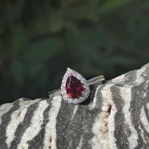 May include: A pear-shaped red gemstone ring with a halo of smaller white gemstones set in silver.
