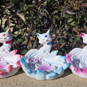 May include: Three ceramic cat figurines with floral bases. Each cat is white with a colored bow tie and a floral design on its head. The bases are in shades of pink, blue, and white, with flower details.
