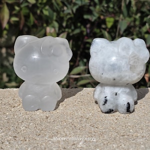 May include: Two small, translucent cat figurines. One is clear, the other is white with black inclusions. The figurines are standing on a textured surface with green foliage in the background. The figurines are in the shape of a popular cartoon character.
