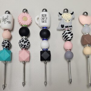 May include: Eight colorful beaded knitting needle tools with various charms, including a cow, a coffee mug, a flower, and a heart. The charms are attached to the top of the needles, which are made of metal.