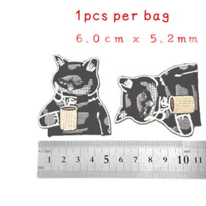 May include: Two black and white embroidered iron-on patches of a cat drinking from a mug. The patches are 6.0 cm by 5.2 mm. The text "1pcs per bag" is written above the patches.