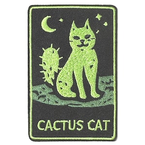 Op de afbeelding: Een rechthoekige geborduurde patch met een limoengroene kat die naast een cactus op een zwarte achtergrond zit. Boven de kat staan een maansikkel en sterren. De tekst CACTUS CAT is onderaan in limoengroen op de patch geborduurd.