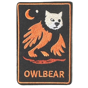 May include: A rectangular embroidered patch with a black background and an orange border. It features an orange crescent moon, a white owl-like head, and an orange winged bear-like body. The word OWLBEAR is stitched in orange at the bottom.