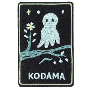 May include: A rectangular embroidered patch with a black background and a light green border. It features a light blue ghost-like figure perched on a tree branch with a white flower, small stars, and the word KODAMA embroidered in white at the bottom.