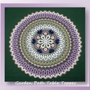 May include: A white crocheted table topper with a green, pink, and purple border. The center of the topper features a floral design with pink and purple flowers and blue butterflies.