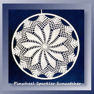 Może przedstawiać: Biały, szydełkowy łapacz słońca z wzorem wiatraka. Łapacz słońca jest zawieszony na obręczy i ma napis "Pinwheel Sparkler Suncatcher" na dole.