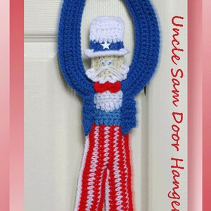 May include: A crocheted Uncle Sam door hanger in blue, white and red. The hanger is shaped like a person with a white hat, a white beard and red and white striped trousers. The text "Uncle Sam Door Hanger" is visible in the image.
