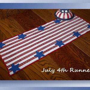 May include: A crocheted table runner in red, white, and blue with blue stars. The runner is on a wooden table and says "July 4th Runner".