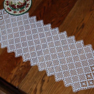 Op de afbeelding: Witte gehaakte tafelloper met een delicaat vierkant patroon. De loper is gemaakt van fijn garen en heeft een kant-achtig, opengewerkt ontwerp.