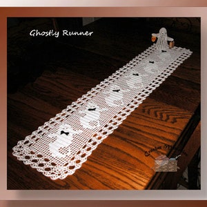 May include: White crocheted table runner with a ghost design. The runner is on a wooden table and has the text "Ghostly Runner" at the top.