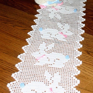 May include: White crocheted table runner with a scalloped edge and a repeating pattern of bunny rabbits. The runner is decorated with pink and blue bows.