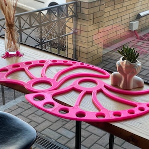 Könnte beinhalten: Hellrosa, fächerförmiges Dekorationsobjekt mit kreisförmigen und ovalen Aussparungen. Es liegt auf einer Holzoberfläche, neben einem kleinen weißen Pflanzgefäß in Kopfform mit einer Sukkulentenpflanze. Eine Glasvase mit getrockneten Pflanzen ist ebenfalls zu sehen.