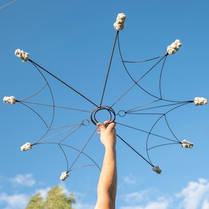 May include: A dark metal fire prop with eight spokes, each tipped with a white, fluffy wick. The prop is held up against a clear blue sky. The central ring is held by a hand.