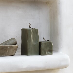 May include: Two olive green block candles with a textured surface and a thin, dark wire detail at the top. The candles are displayed on a white shelf, with a mortar and pestle visible on the left. The text "RAW BLOCK | SET OF 2" is at the bottom.