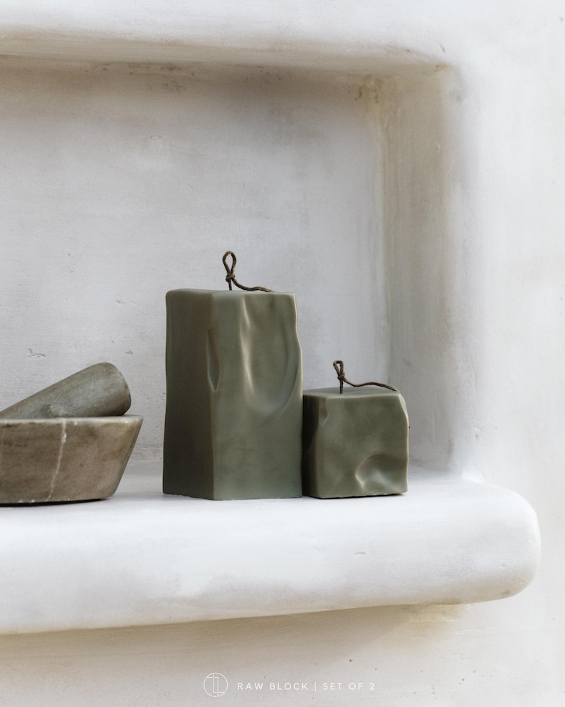 May include: Two olive green block candles with a textured surface and a thin, dark wire detail at the top. The candles are displayed on a white shelf, with a mortar and pestle visible on the left. The text "RAW BLOCK | SET OF 2" is at the bottom.