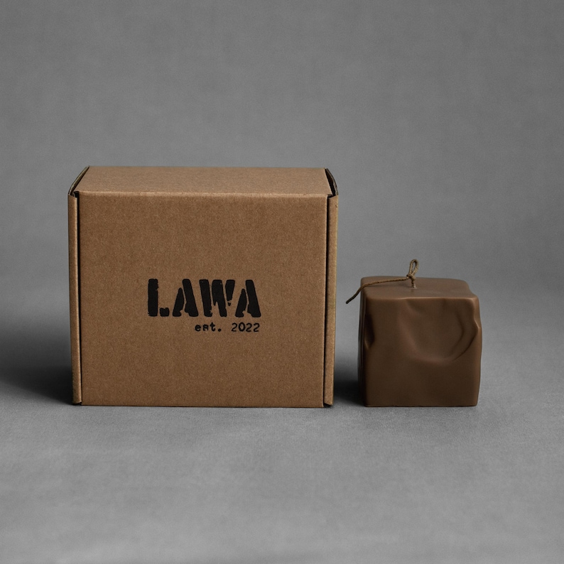 May include: A brown square candle with a textured surface and a small twine detail, next to a brown cardboard box. The box has the word "LAWA" and "ent. 2022" printed on it in black. The background is a solid gray.