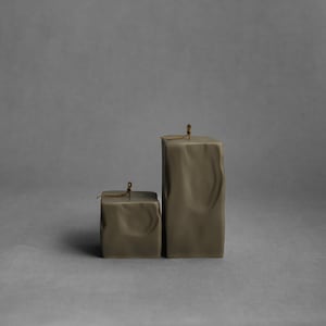 May include: Two olive-green square candles of different sizes. The candles have a textured, wrinkled appearance and a small wick at the top. The background is a solid gray.