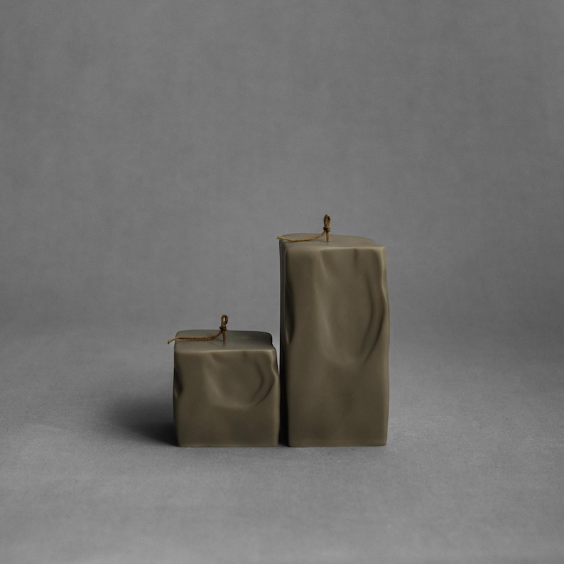 May include: Two olive-green square candles of different sizes. The candles have a textured, wrinkled appearance and a small wick at the top. The background is a solid gray.