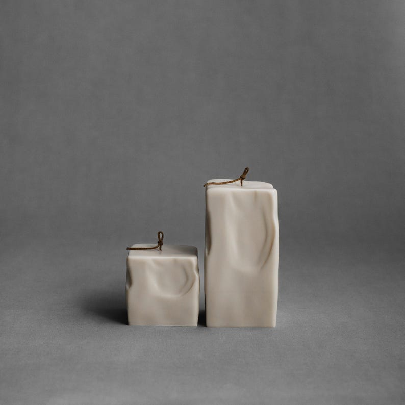 May include: Two off-white, square candles with a marbled effect. The candles have a textured surface and are topped with a small, brown string tied in a bow. One candle is shorter and wider than the other.