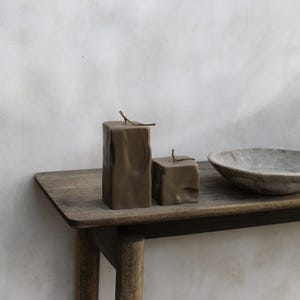 May include: Two brown, textured block candles with a twig on top, displayed on a wooden table. A gray, round bowl is also on the table. The candles are part of a set of two.