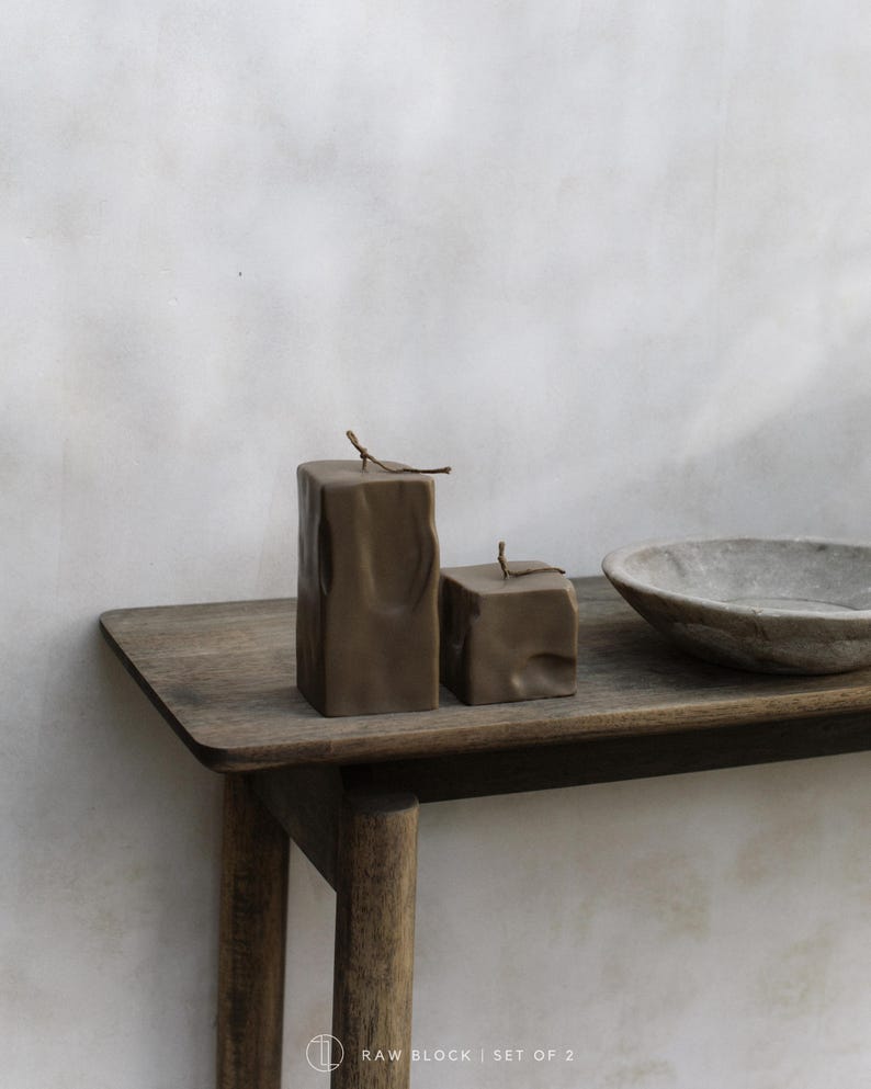 May include: Two brown, textured block candles with a twig on top, displayed on a wooden table. A gray, round bowl is also on the table. The candles are part of a set of two.