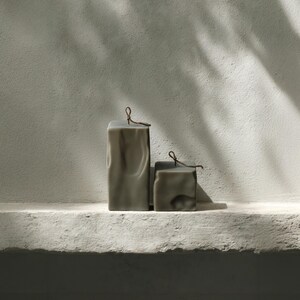 May include: Two earthy green block candles with a textured surface and a small loop at the top. The candles are different sizes and sit on a white ledge against a textured wall. The text at the bottom reads "RAW BLOCK SET OF 2 | EARTHY GREEN".