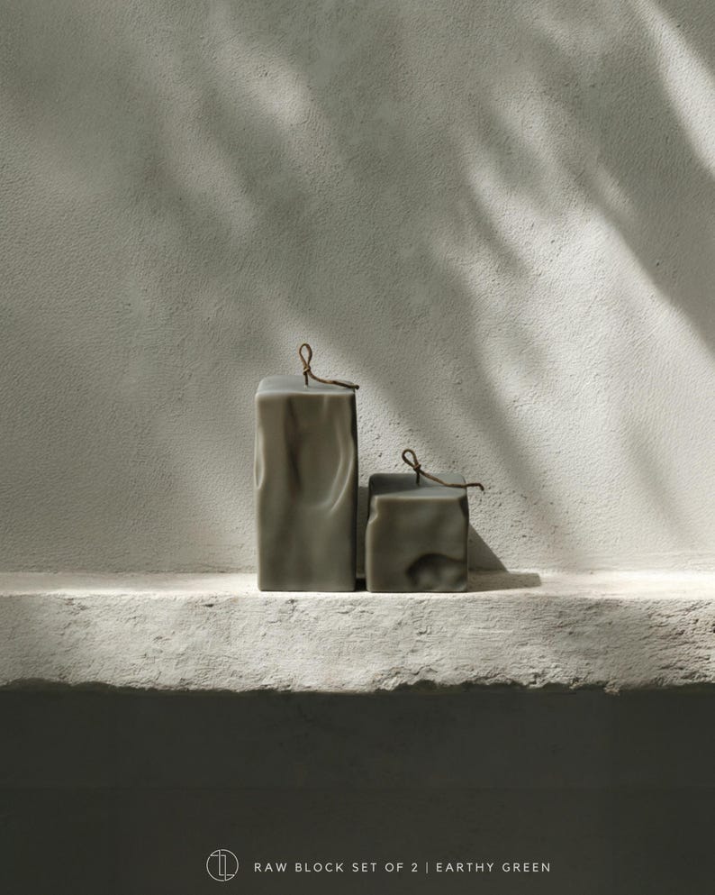 May include: Two earthy green block candles with a textured surface and a small loop at the top. The candles are different sizes and sit on a white ledge against a textured wall. The text at the bottom reads "RAW BLOCK SET OF 2 | EARTHY GREEN".