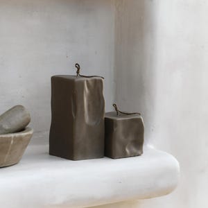 May include: Two brown block candles with a textured surface and a small loop at the top. The candles are displayed on a white shelf. The text "RAW BLOCK | SET OF 2" is at the bottom.