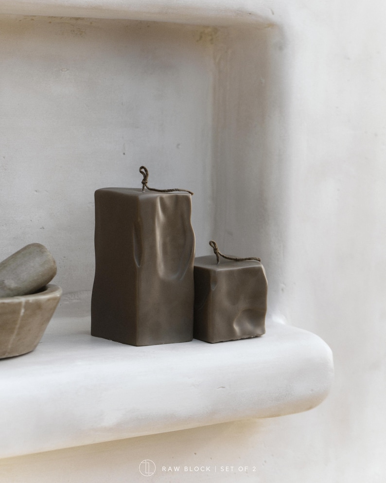 May include: Two brown block candles with a textured surface and a small loop at the top. The candles are displayed on a white shelf. The text "RAW BLOCK | SET OF 2" is at the bottom.