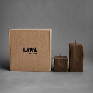 May include: A brown cardboard box with the text "LAWA est. 2022" printed in black. Two brown square candles with a textured surface and a small string tied at the top are next to the box.