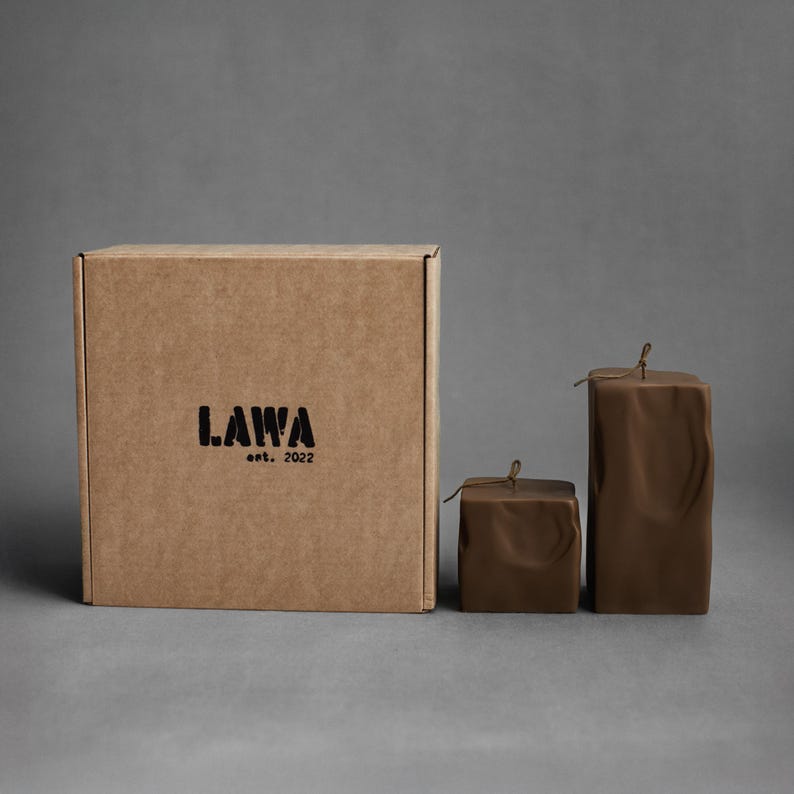 May include: A brown cardboard box with the text "LAWA est. 2022" printed in black. Two brown square candles with a textured surface and a small string tied at the top are next to the box.