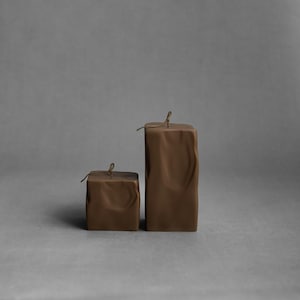 May include: Two brown square candles with textured surfaces and a string tied around the top. The candles are different sizes, with the larger one standing upright and the smaller one beside it. The background is a solid gray.