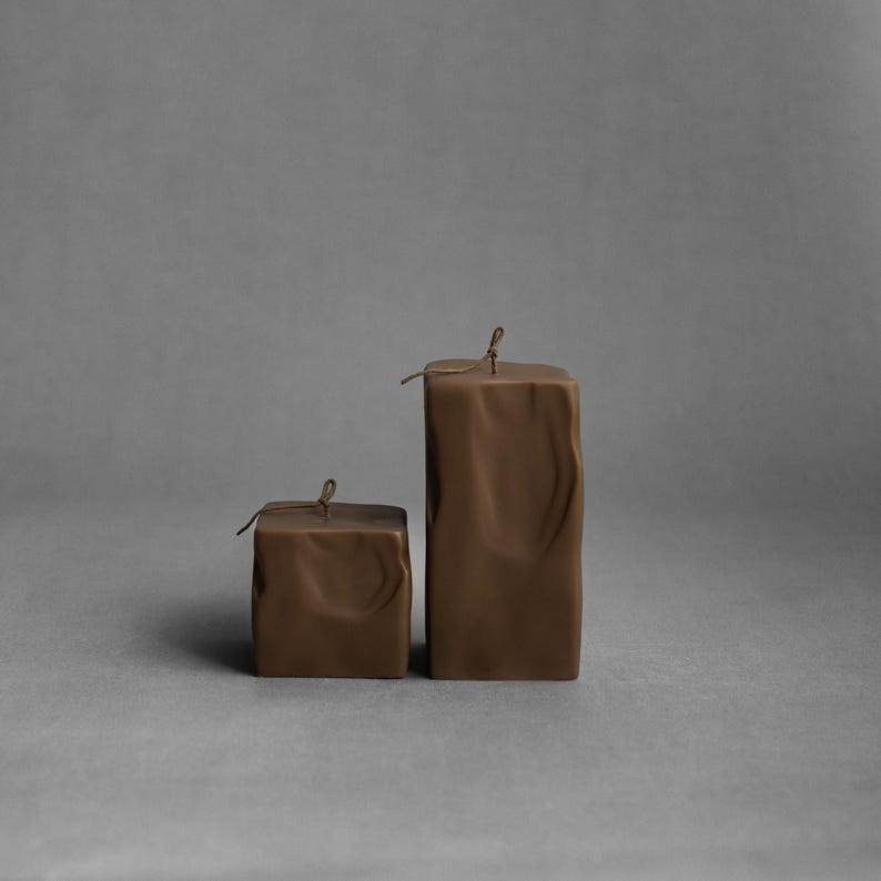 May include: Two brown square candles with textured surfaces and a string tied around the top. The candles are different sizes, with the larger one standing upright and the smaller one beside it. The background is a solid gray.