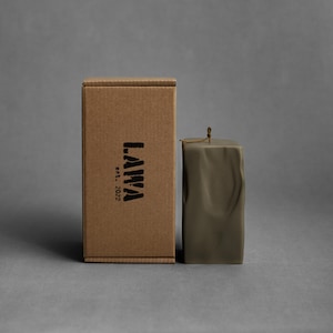 May include: A square, olive-green candle with a textured surface stands next to a brown cardboard box. The box has the word "LAMA" printed on it in black, along with "est. 2022". The candle has a small, decorative string at the top.