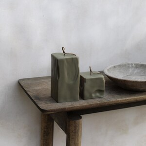 May include: Two olive green, square-shaped candles with a textured surface and a small wick. The candles are displayed on a wooden table next to a gray bowl. The text "RAW BLOCK | SET OF 2" is at the bottom.