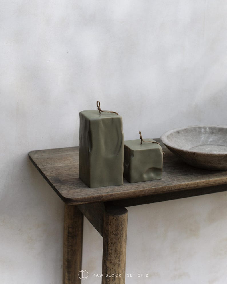 May include: Two olive green, square-shaped candles with a textured surface and a small wick. The candles are displayed on a wooden table next to a gray bowl. The text "RAW BLOCK | SET OF 2" is at the bottom.