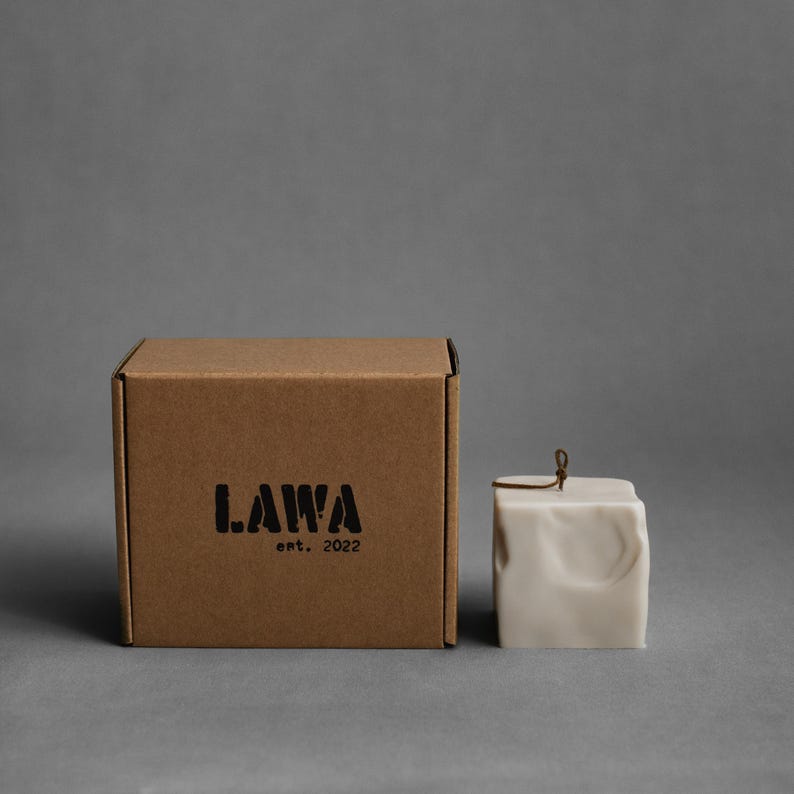 May include: A square, off-white candle with a textured surface and a small metal loop on top. Beside it is a brown cardboard box with the word "LAWA" and "ent. 2022" printed in black.