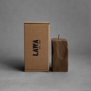 May include: A brown square candle with a textured surface and a wick, next to a cardboard box. The box has the word "LAWA" printed on it, with "est. 2022" below it.